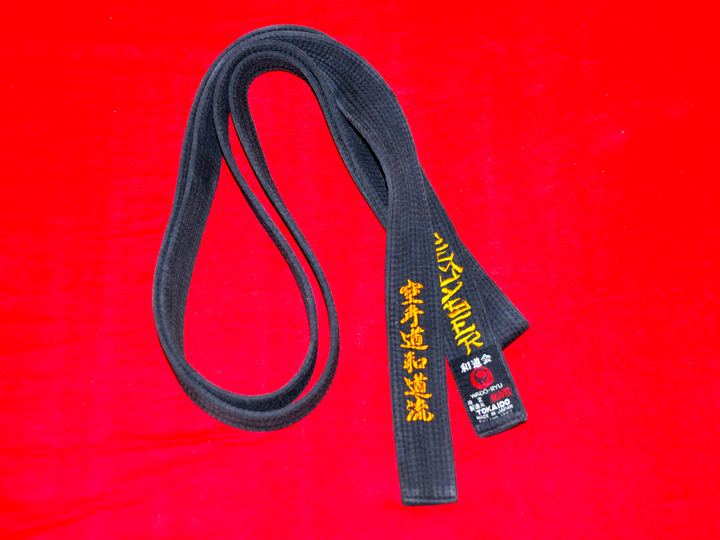 A black karate belt with gold embroidered lettering on a red background